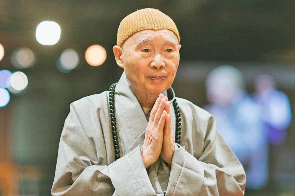 Master Chin Kung biography shown through an image of the revered Pure Land Buddhism teacher in traditional robes, with prayer beads and hands in respectful greeting.